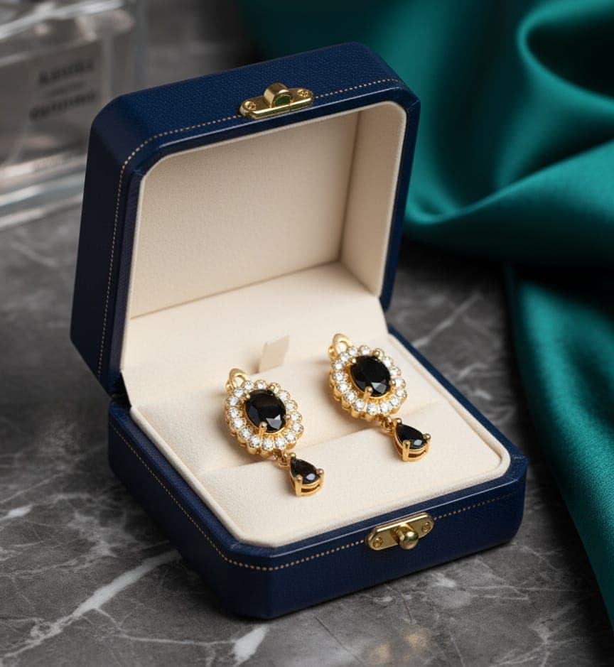 A delicate gold-plated earring featuring a golden flower design studded with shiny stones, a luxurious black stone in the middle, and a luxurious black teardrop stone hanging from it. Clip-Ons